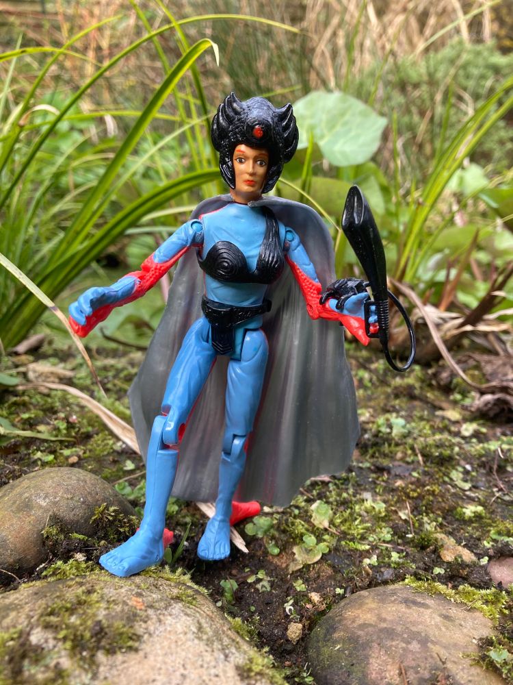 Power Lords Shaya - a regal woman in a blue bodysuit, ornate crown weird black bikini and silver cape. Has a strange weapon/microphone 