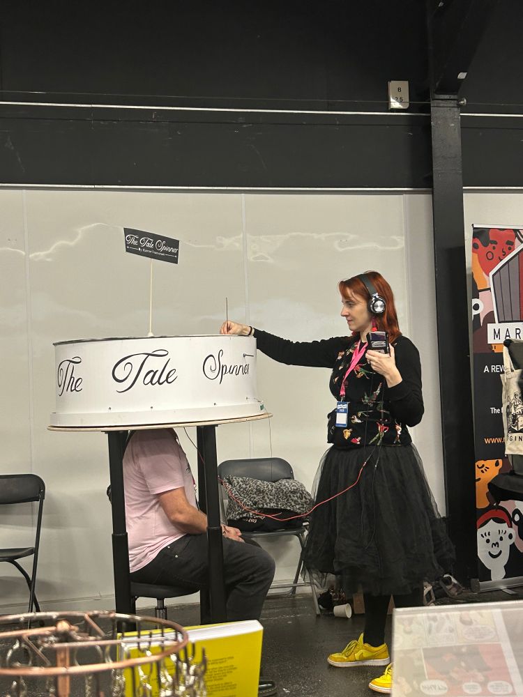 A person sits on a stool with their head inside a big circular structure. Karrie, wearing headphones and holding a Walkman style device, gently adjusts bolts and spins the wheel around. She is wearing an embroidered velvet waistcoat, black tutu and yellow trainers.
