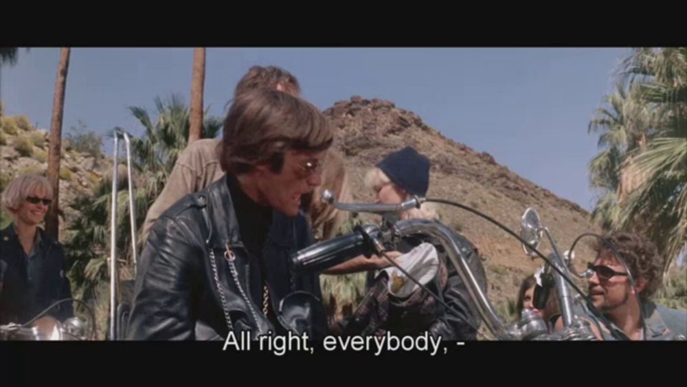 a man in a leather jacket says " all right everybody " while riding a motorcycle