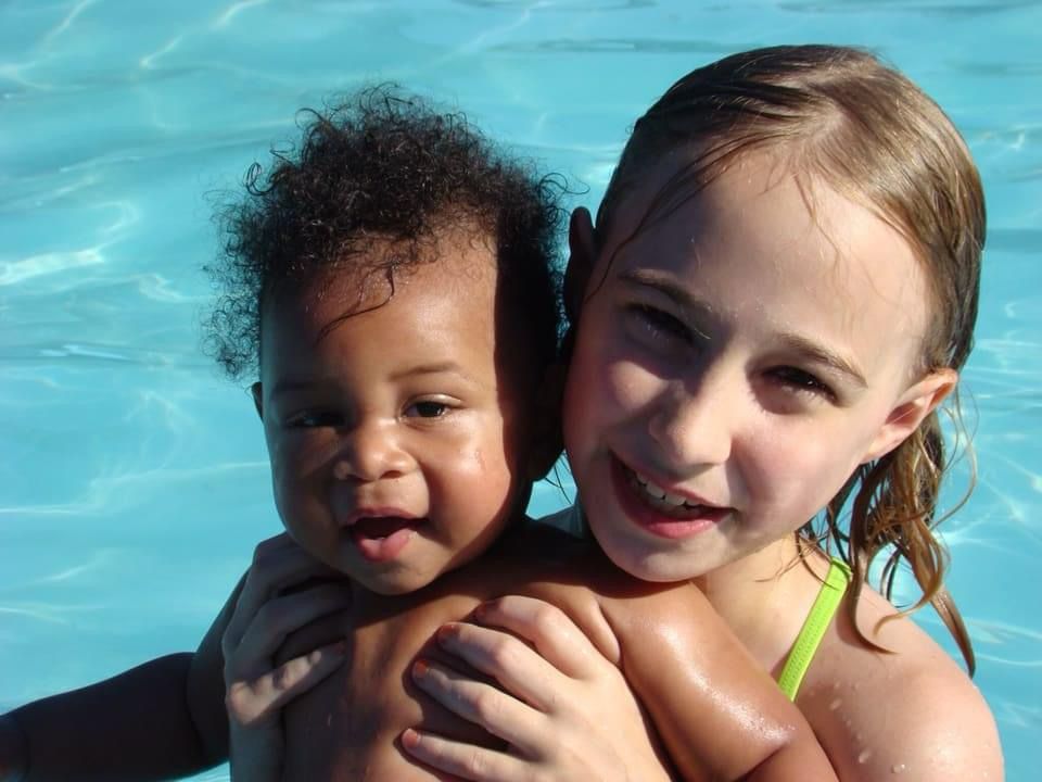 A brown-skinned baby is held up by a young blonde girl in a swimming pool. Bright sunlight shines from their left, casting shadows on the other side of their faces. The water is a bright aqua color. There are faint ripples in the water.  