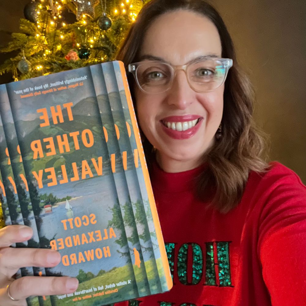 A woman with dark shoulder length hair, and a red Christmas jumper holds up the book The other valley by Scott Alexander Howard