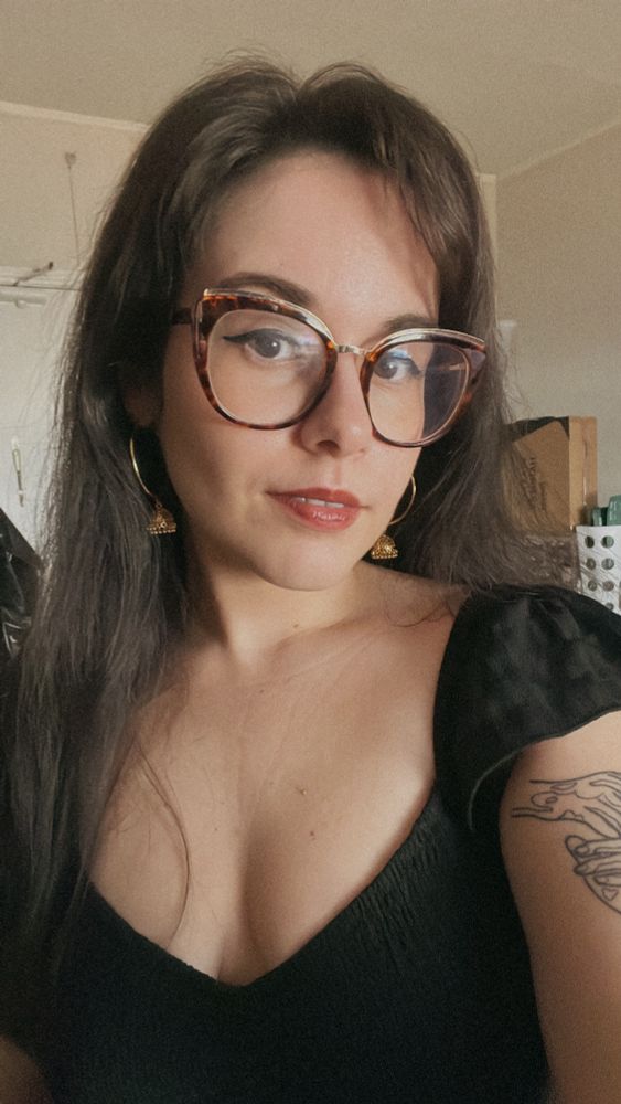 selfie of Andrea wearing a black dress. her hair is long and down. she has brown glasses on and has a full face of makeup on.  her head is a bit tilted to the side and she looking at the camera with a slight smirk. 