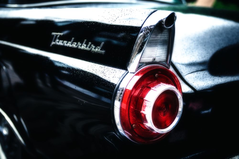 A close-up view of a classic Ford Thunderbird's tail fin and round red taillight is prominently displayed, with the word "Thunderbird" elegantly scripted on the shiny black surface. Raindrops add a subtle sheen, enhancing the vintage feel of the vehicle.