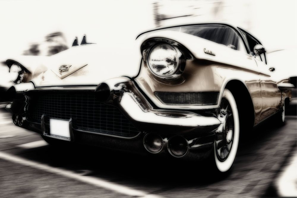 A classic Cadillac Coupe DeVille with a prominent grille and shiny chrome details is parked outdoors, showcasing its vintage aesthetic. The photograph has a slightly blurred effect, enhancing the nostalgic feel of the scene.