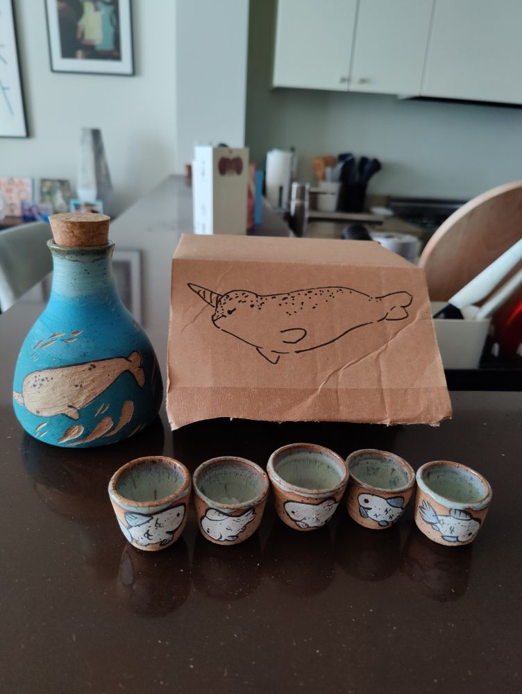 A sake set consisting of a pitcher and five glasses sit on a countertop next to a cardboard box. The set is ceramic; the pitcher features a narwhal and the cups have fish hand drawn on them. 