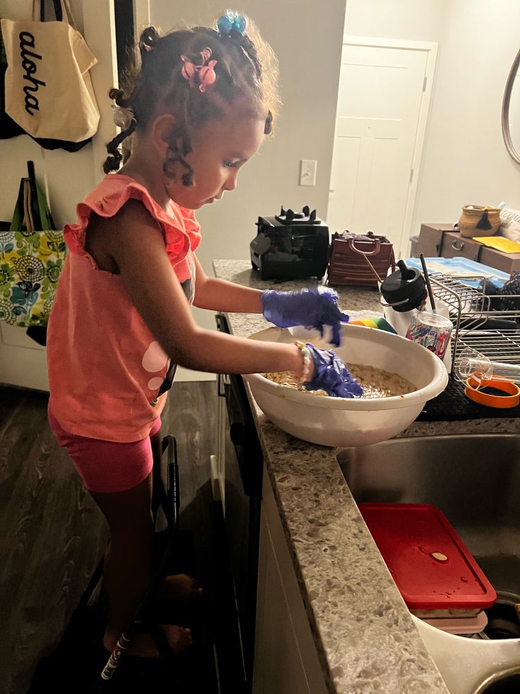 Little girl wearing pink standing on a step stool with purple disposable gloves rinsing pumpkin seeds in a bowl.