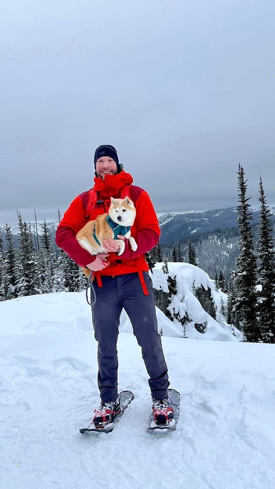 A man holding a dog while standing on a snow covered peak, wearing snow shoes