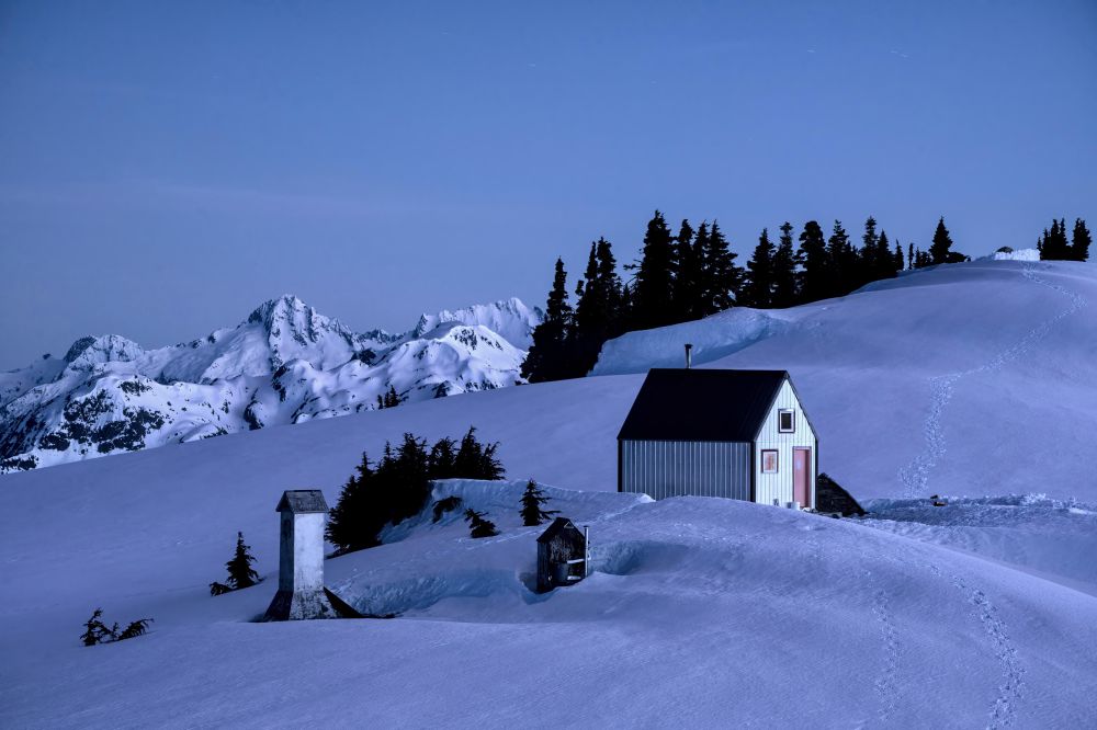 A solitary cabin with a dark roof stands in a snowy landscape, surrounded by mountains and pine trees under a twilight sky. Snow covers the ground, and a small, weathered structure is nearby.