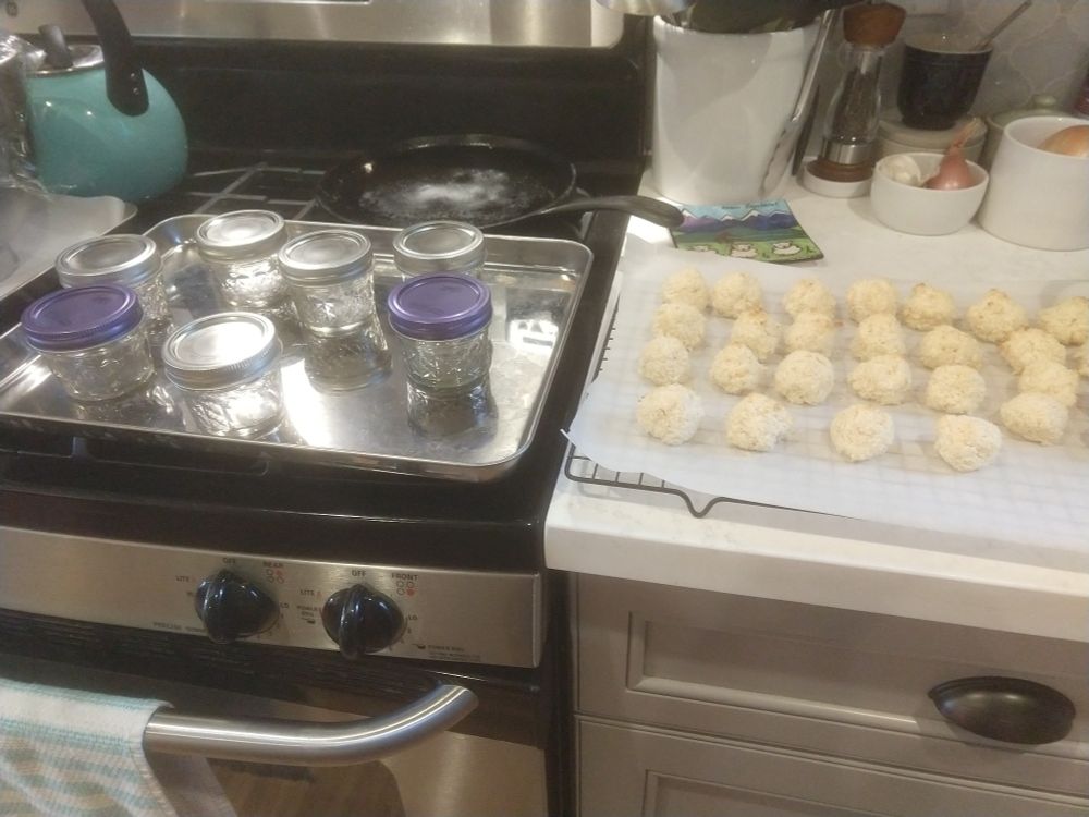 Tray with seven small glass jars sitting on the stove with a batch of coconut macaroons on a cooling rack to the left.