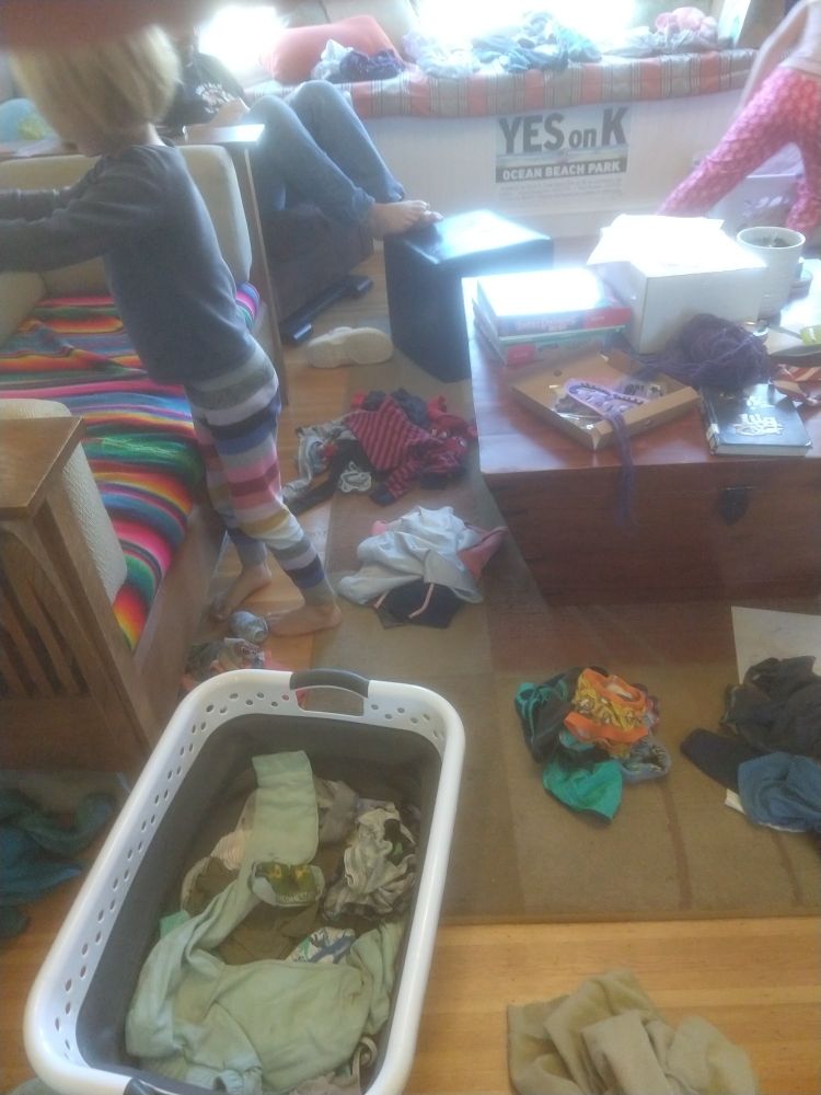 An image of a small child near a laundry basket with piles of sorted clothes all around. 