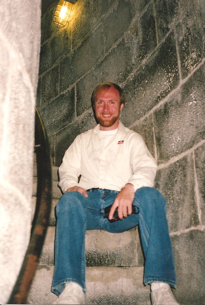 Young bearded man in blue jeans and a white shirt sitting on stone steps of a lighthouse's internal spiral staircase.