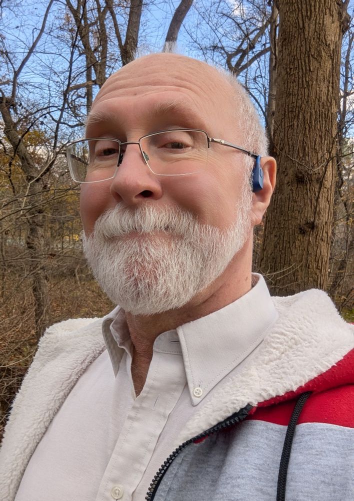 Old guy in a fluffy hoodie out walking in a wooded municipal park.