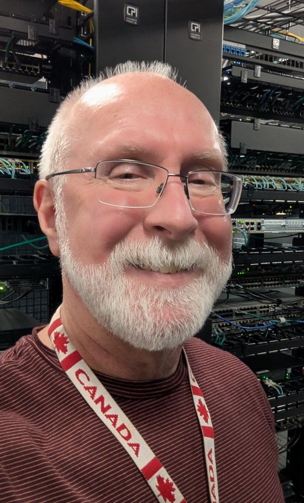 Old guy in glasses and red shirt standing in front of a row of network racks in a data center.