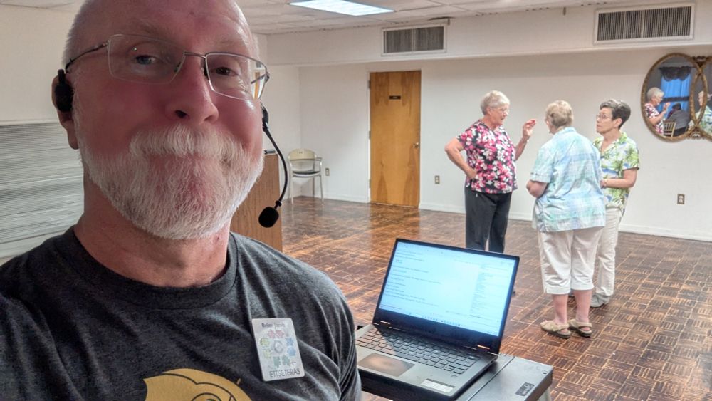 Old guy wearing a microphone headset in front of a laptop computer. A dance floor with three dancers is in the background.