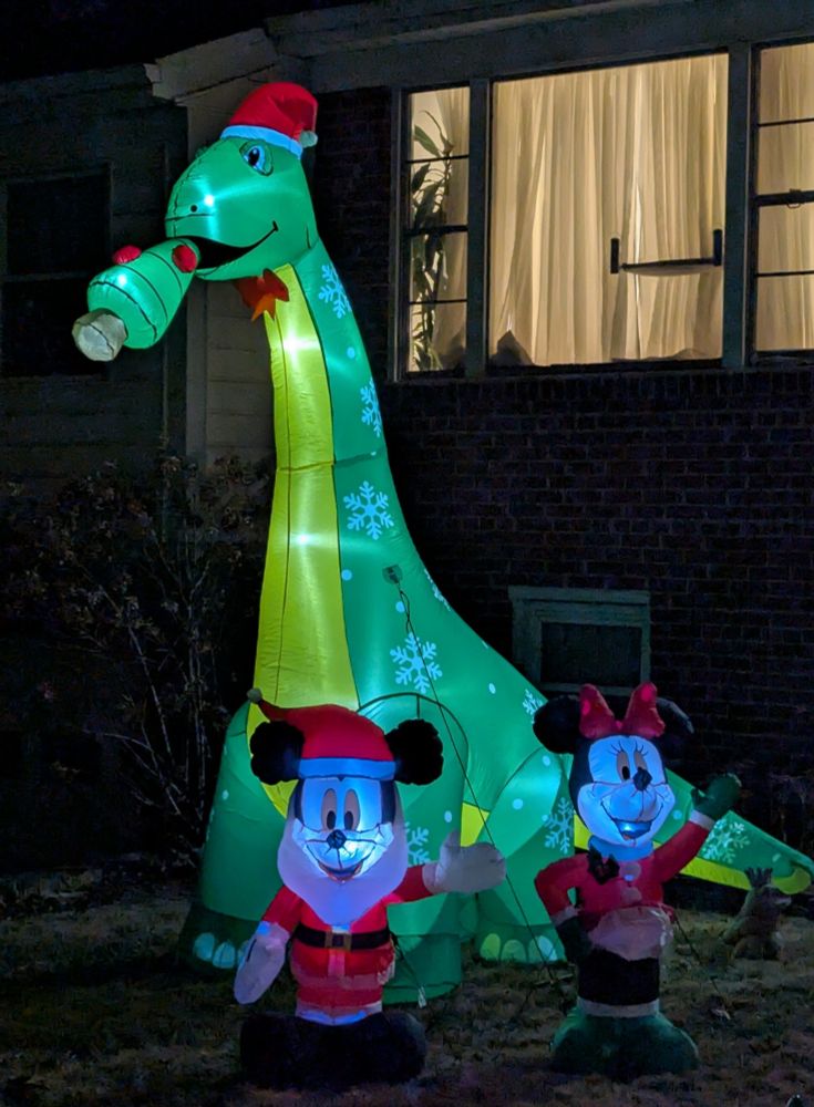 Inflatable lawn Xmas decoration of a large sauropod dinosaur wearing a red toque and eating an Xmas tree.