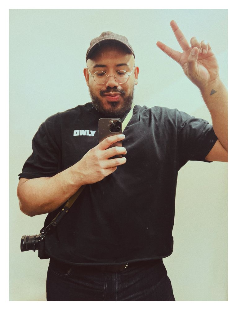 marvin gesturing the peace sign in a cap, black tee shirt and jeans with a Nikon Zf on a crossbody strap. 