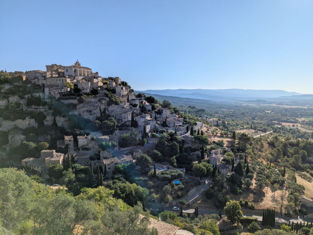 A photograph of the majestic Provençal village of Gordes, brown houses climbing up a hillside with green wine country beyond.