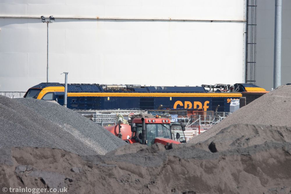 a large blue and yellow locomotive obstructed by some stones and a red tractor. In the background are giant white fuel storage silos.