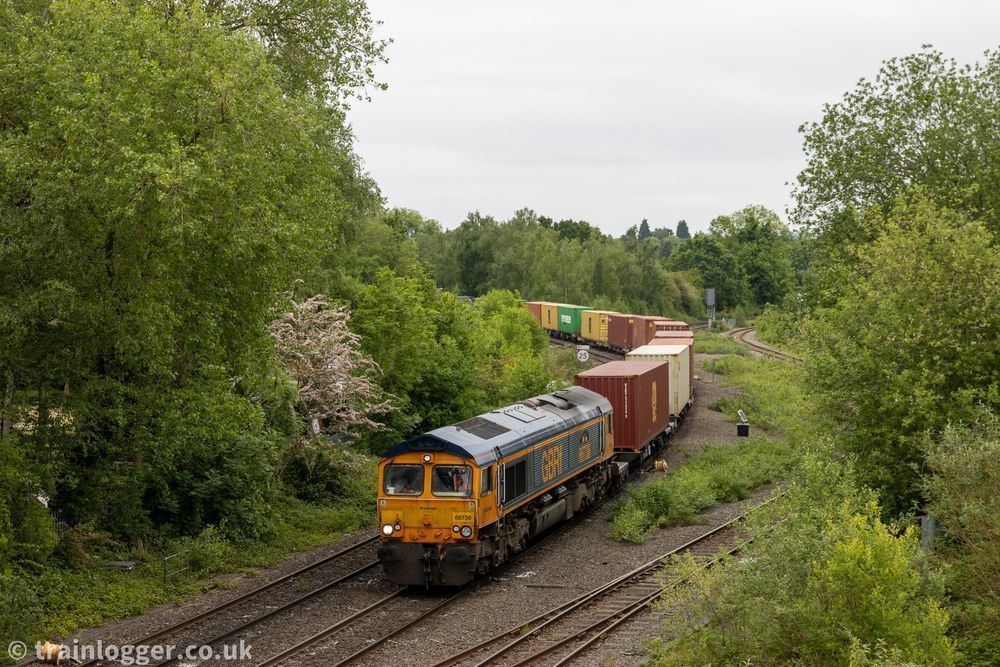 a dark blue & yellow railway locomotive is seen hauling some wagons loaded with container boxes. It is passing through a leafy scene