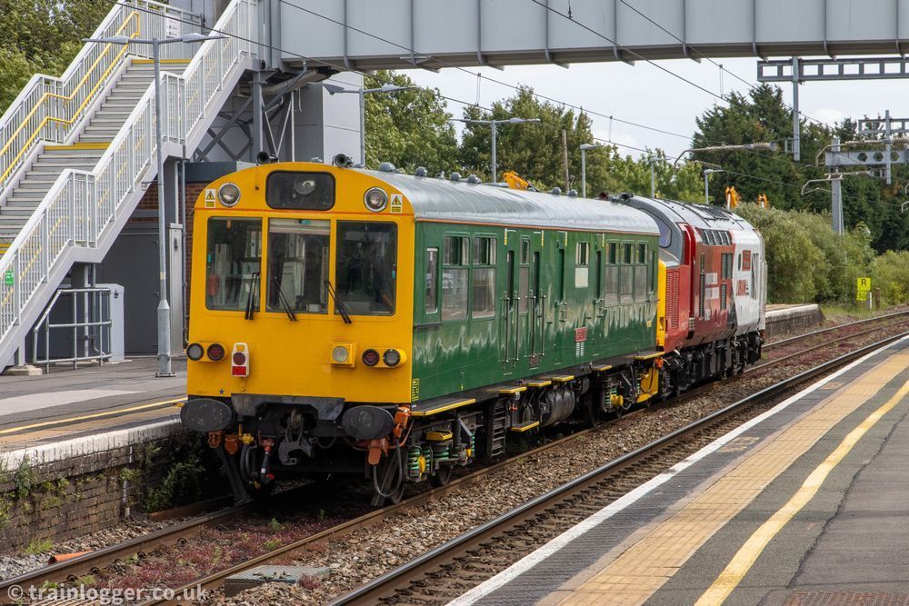 a green carriage is being hauled by a smart white, red, grey and yellow locomotive going under a bridge