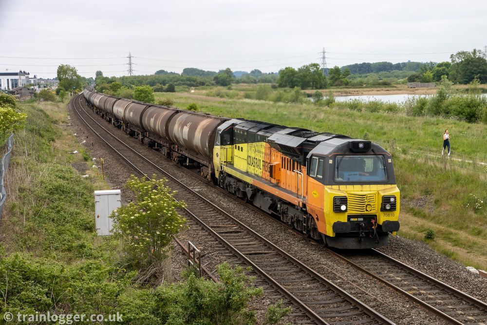 a yellow, black & orange locomotive is seen hauling some cylindrical tanker wagons through a green landscape. In the background is a lake, some pylons, a jogger and some industrial units
