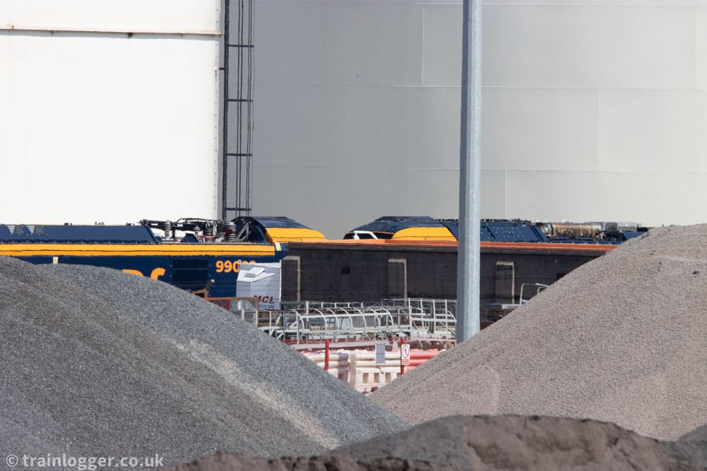 two large blue and yellow locomotive obstructed by some stones and portacabins.  In the background are giant white fuel storage silos.