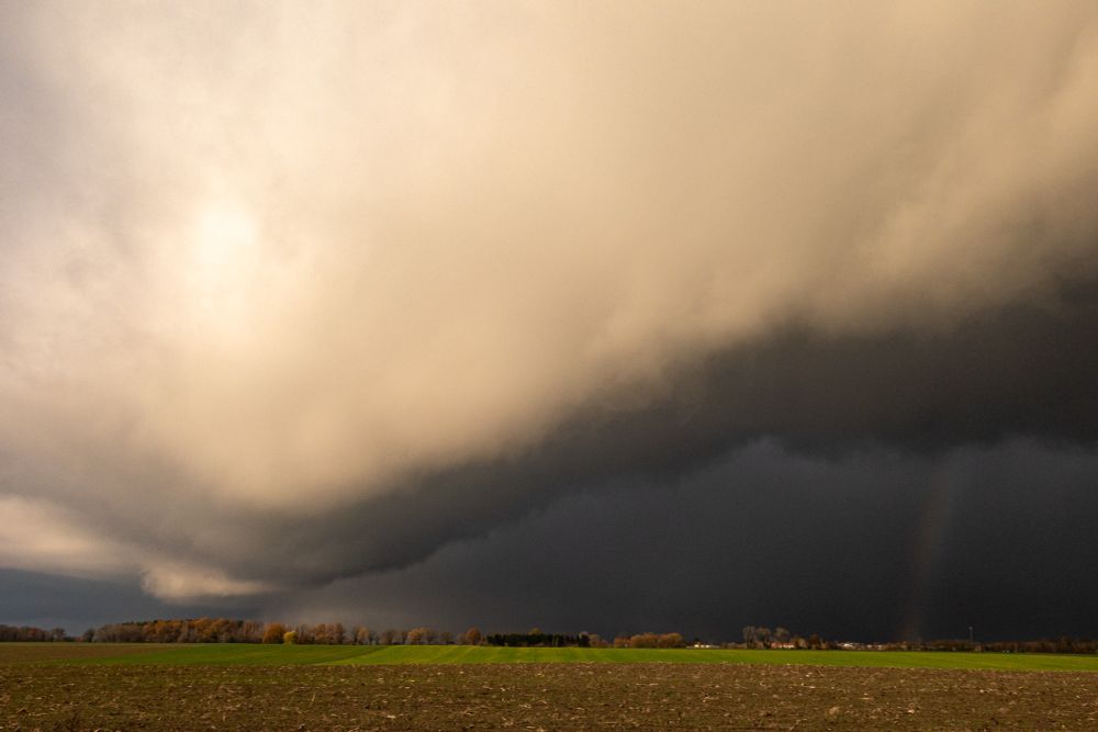 Das Foto zeigt eine weitläufige Landschaft mit Feldern im Vordergrund und einem ausgedehnten dunklen Waldstreifen in der Distanz. Darüber dominiert ein dramatischer Himmel: eine massive, tiefhängende Wolkenfront zieht von links nach rechts ins Bild und bildet einen deutlichen Kontrast zwischen hellen, warmen Wolkentönen und einem nahezu schwarzen Kernbereich. Rechts im Bild ist ein schwacher Regenbogen zu erkennen, der aus der dunklen Wolkenzone austritt. Die Szene vermittelt Wetterumschwung, vermutlich kurz vor oder nach einem starken Regenschauer.
