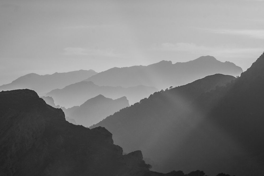 Schwarzweißaufnahme einer Berglandschaft mit mehreren hintereinanderliegenden Bergketten, die sich in weichen Abstufungen von hell nach dunkel in die Ferne ziehen. Zwischen den Hängen fällt schräg ein Sonnenstrahl durch den leichten Dunst und verstärkt die Tiefe des Bildes. Die Szenerie wirkt ruhig, zeitlos und minimalistisch.