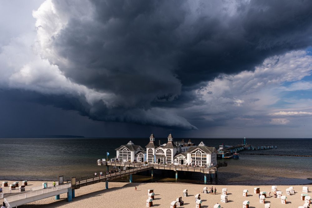 Eine dramatische Gewitterfront zieht über die Seebrücke von Sellin an der Ostsee. Dunkle, bedrohliche Wolkenwalzen rollen über das Meer, während der Strand mit Strandkörben und vereinzelten Menschen noch im Licht liegt.