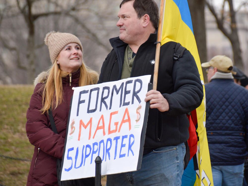A pro- Jan 6th beige male(black jacket & olive colored henley shirt) and woman (beige beanie and maroon jacket). Stand side by holding a sign that reads "FORMER $MAGA$ SUPPORTER" & a Ukraine flag. It is unclear who the former MAGA supporter is.