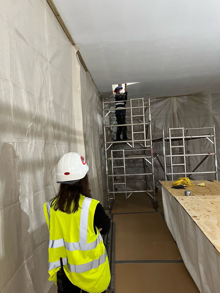 The project manager, in a hi vis jacket and hard hat stands in an room emptied of furniture, the walls and floor protected, watching as a contractor on a portable scaffold tower removes a piece of the ceiling above them.