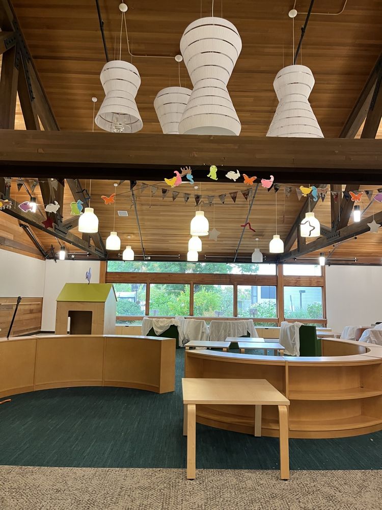 A wood paneled room with a wall of windows, a tall ceiling, and white hanging lamps. An S-shaped bookshelf bisects the room and there is a children's playhouse with a green roof on the left. 