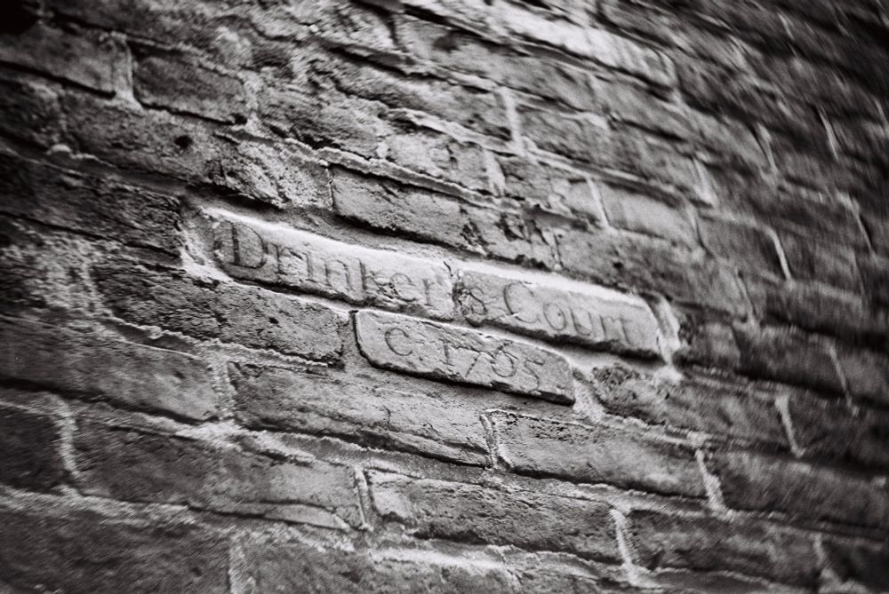 close black and white photo of a rough brick wall with "Drinker's Court / C. 1765" carved on smoother bricks in the center; there is a slightly swirling vignette effect