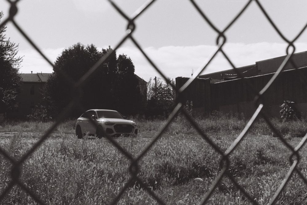 black and white photo of a grassy empty lot through a chainlink fence; a shiny white car is parked in the middle of the lot