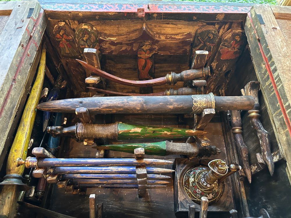 a photo of the interior of an antique Jinn warfare case with painted spikes, an enamel jug, and what appear to be wooden scimitars