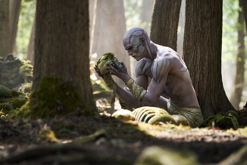 A bald man with different patches of stitched up skin squats down in a forest next to a skeleton as he gazes at it's skull in the morning light.