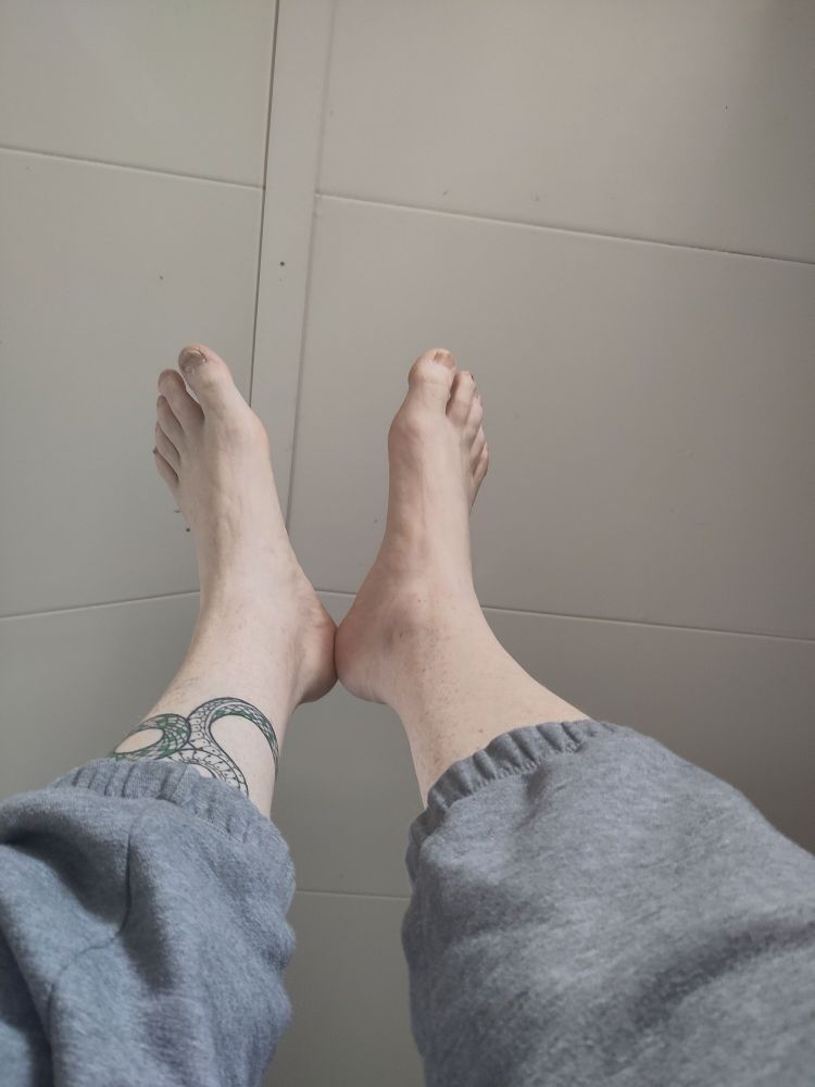 Foto van mijn onderbenen en voeten, op mijn rug liggend genomen. Je ziet dat ik een grijze joggingbroek aan heb en een slangen tattoo op mijn linker onderbeen.