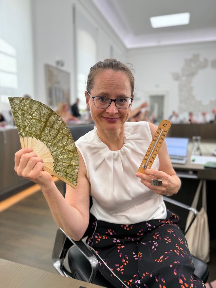 Eine Frau sitzt in einem modernen Sitzungssaal auf einem Bürostuhl und lächelt in die Kamera. Sie trägt eine weiße ärmellose Bluse und einen dunklen Rock mit buntem Muster. In ihrer rechten Hand hält sie einen Fächer mit grün-goldenem Muster, in der linken ein Holzthermometer, das rund 30 Grad anzeigt. Im Hintergrund sind weitere Personen an Tischen zu sehen, offenbar in einer Sitzung oder Versammlung.