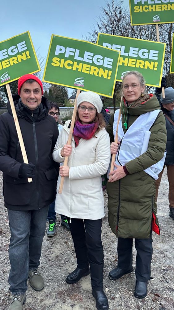 Ein Mann und zwei Frauen in Winterjacken mit Schildern, auf denen zu lesen ist: Pflege sichern