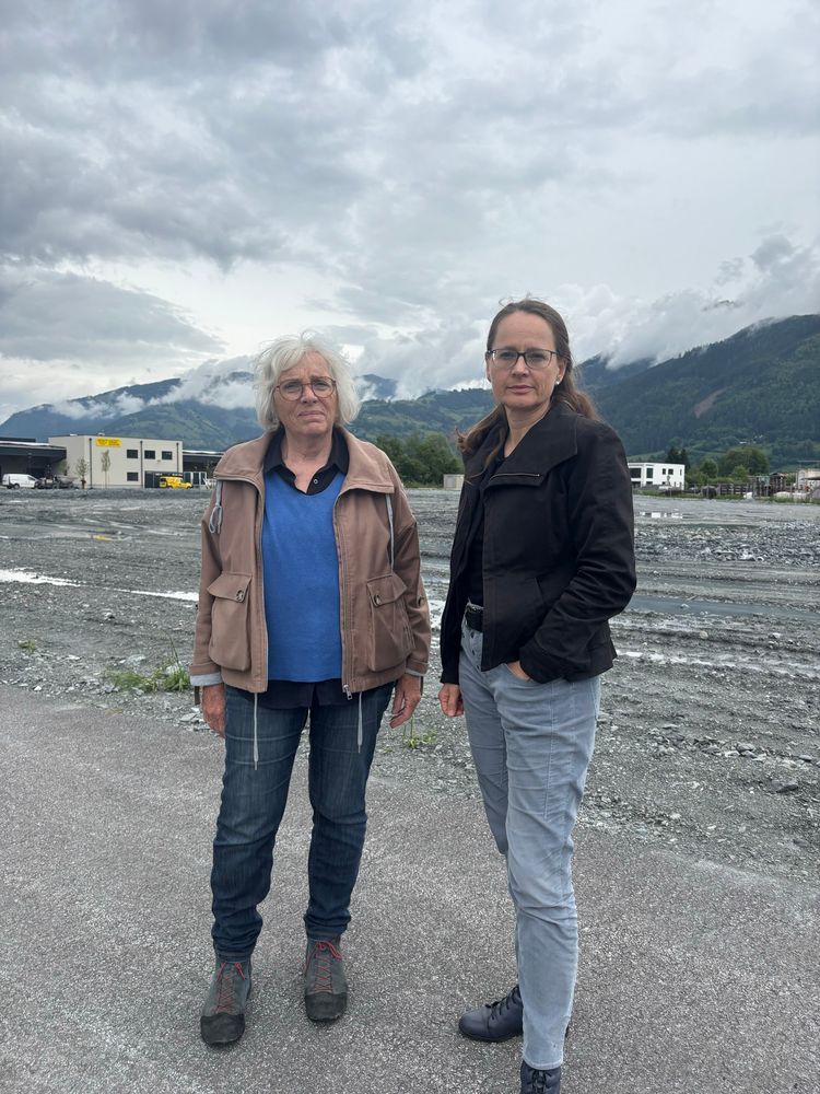 Zwei Frauen stehen auf einer asphaltierten Fläche vor einem großflächigen, steinigen Gelände mit Baustellencharakter. Im Hintergrund sind Industriegebäude und eine bewaldete Berglandschaft mit tief hängenden Wolken zu sehen. Beide Frauen blicken ernst in die Kamera, tragen wetterfeste Kleidung und Brillen.