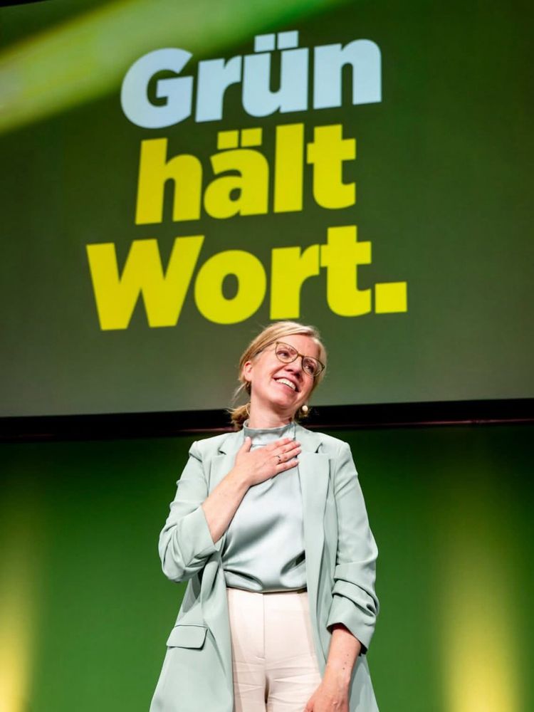 Eine Frau steht lächelnd auf einer Bühne vor einem grünen Hintergrund mit der Aufschrift „Grün hält Wort.“ in großen Buchstaben. Sie trägt eine mintgrüne Jacke, ein helles Oberteil und eine helle Hose. Eine Hand liegt auf ihrer Brust, sie wirkt gerührt und dankbar. Die Stimmung ist positiv und zugewandt.