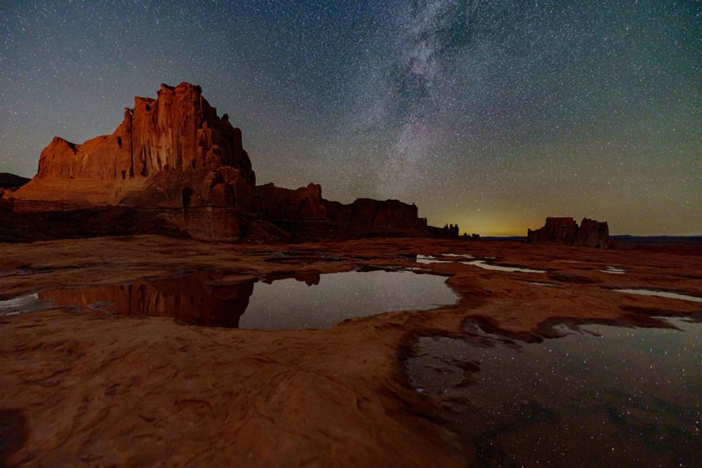 A photo of a reddish landscape with a starry sky and the Milky Way stretching vertically near the center of the sky. In the foreground are several pools of water reflecting the starry sky.