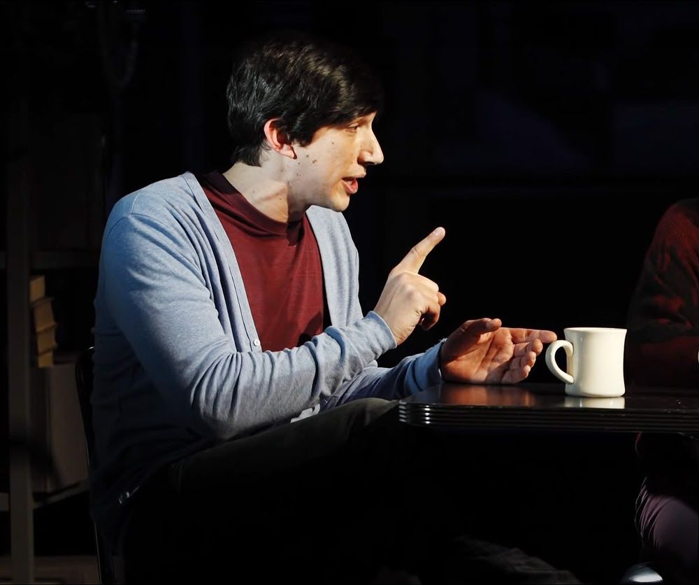 Adam Driver as Louis Ironson in Angels in America, pointing a finger and speaking animatedly over a cup of coffee