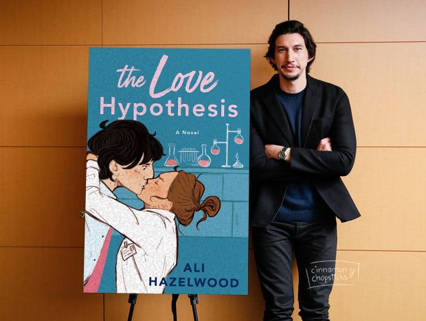 A manip of an Adam Driver photo picturing him standing next to a poster of the cover for The Love Hypothesis by Ali Hazelwood