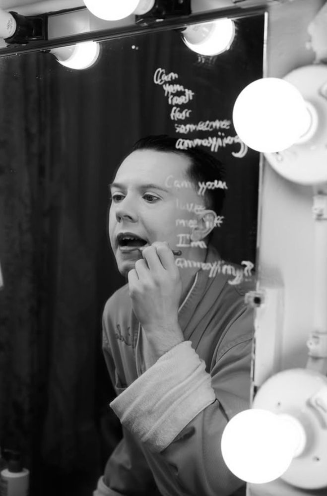 Black-and-white portrait of Cole Escola in a robe applying lip liner in an old-fashioned lighted mirror with the bulbs around the frame. Written on the mirror beside and slightly overlapping with them: “Can you root for someone annoying? Can you love me if I’m annoying?”