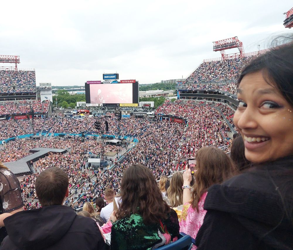 Me at the eras tour up in the nosebleeds smiling at the camera. I have tan skin, dark hair and eyes, slightly crooked teeth and sparkly makeup. Wearing a black hoodie.