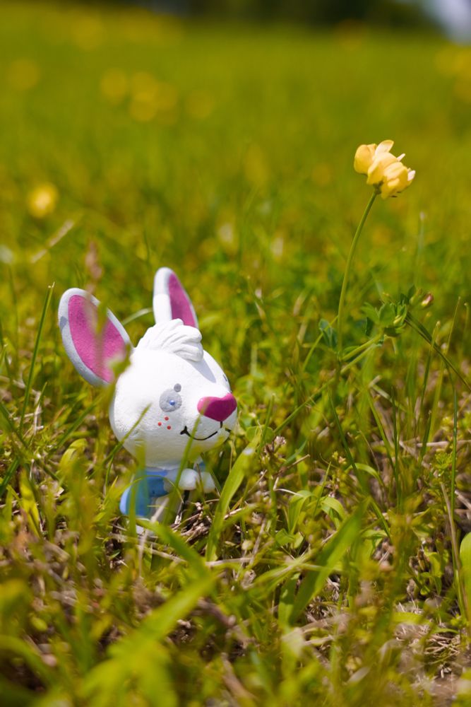 A figurine of Lea, a white, blue, and purple anthropomorphic rabbit, sitting in the grass next to a yellow flower. 