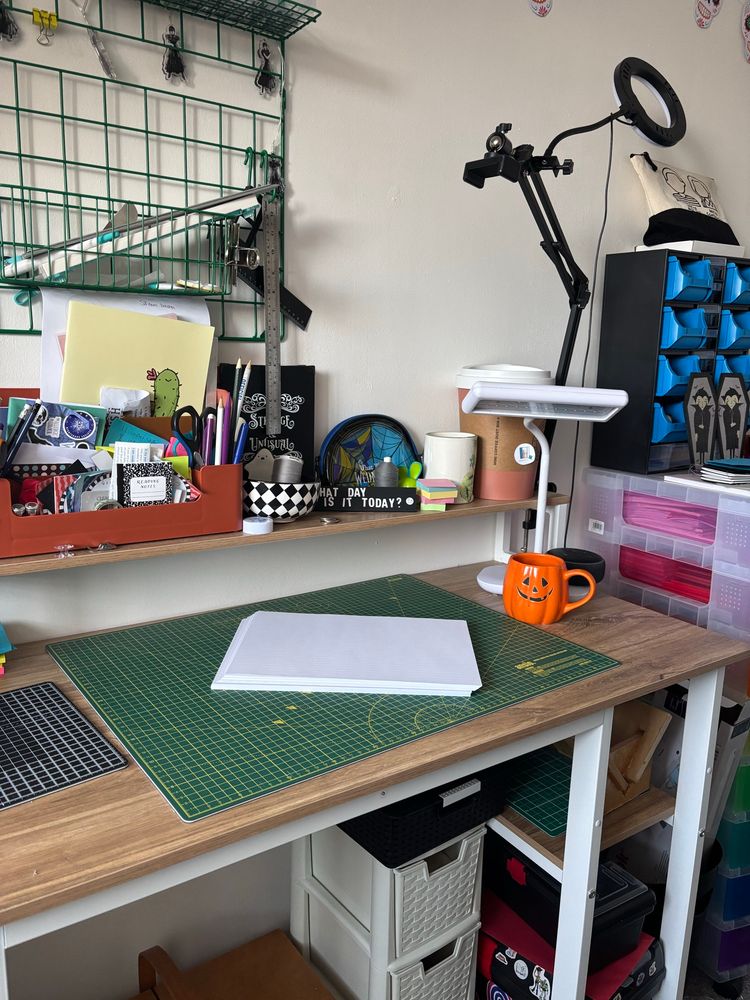 My desk in my craft room with a cutting mat, a pile of paper, a pumpkin mug of coffee and a desk tidy that really could do with a tidy up 