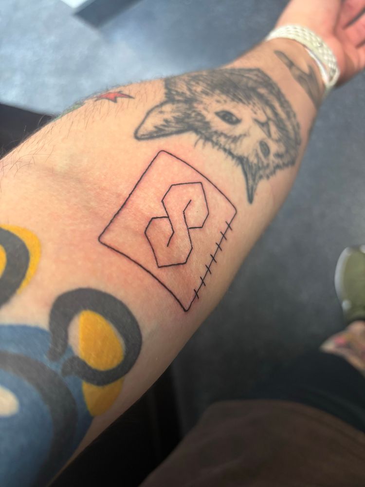 A photo of a forearm with tattoos, with a new tattoo of the “cool S” inside a notebook page