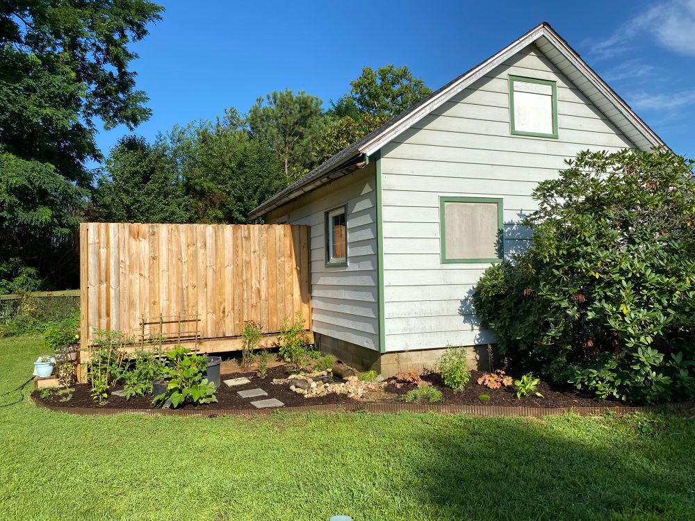 Picture of my beautiful cottage with the deck and the fence and the new flowerbeds almost entirely mulched with nice brown mulch, and all the plants, vegetables, herbs, and flowers tucked into it. You can also see the new gray concrete square pavers laid into the beds.